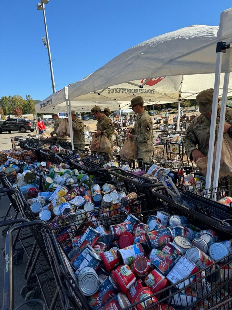 A handful of service members in camouflage uniforms stand outside near a group of large baskets filled with canned goods.