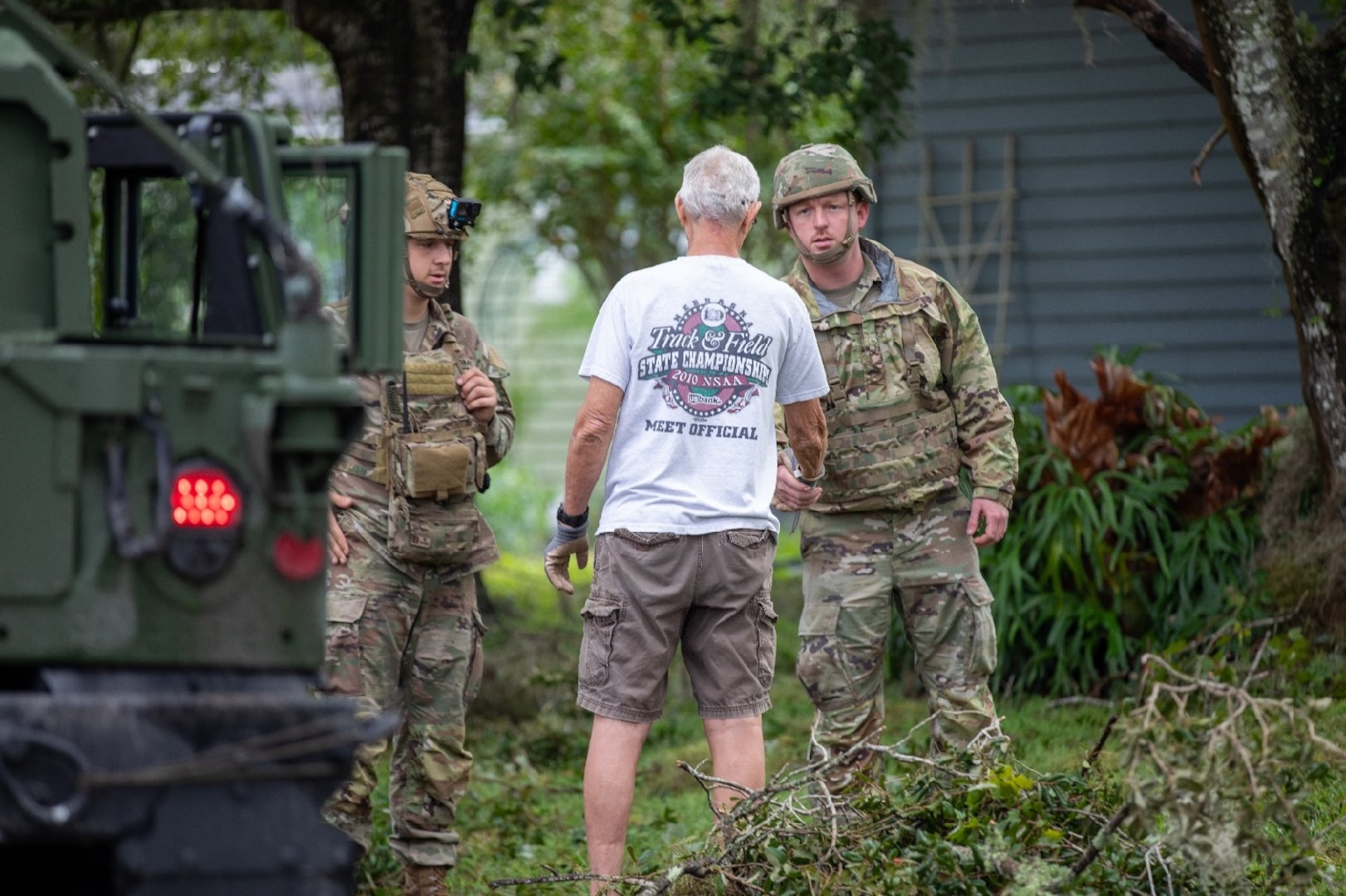 Two service members in camouflage uniforms and a civilian man in shorts and a T-shirt stand outside near a military vehicle amid foliage and debris. One of the service members is shaking hands with the civilian.