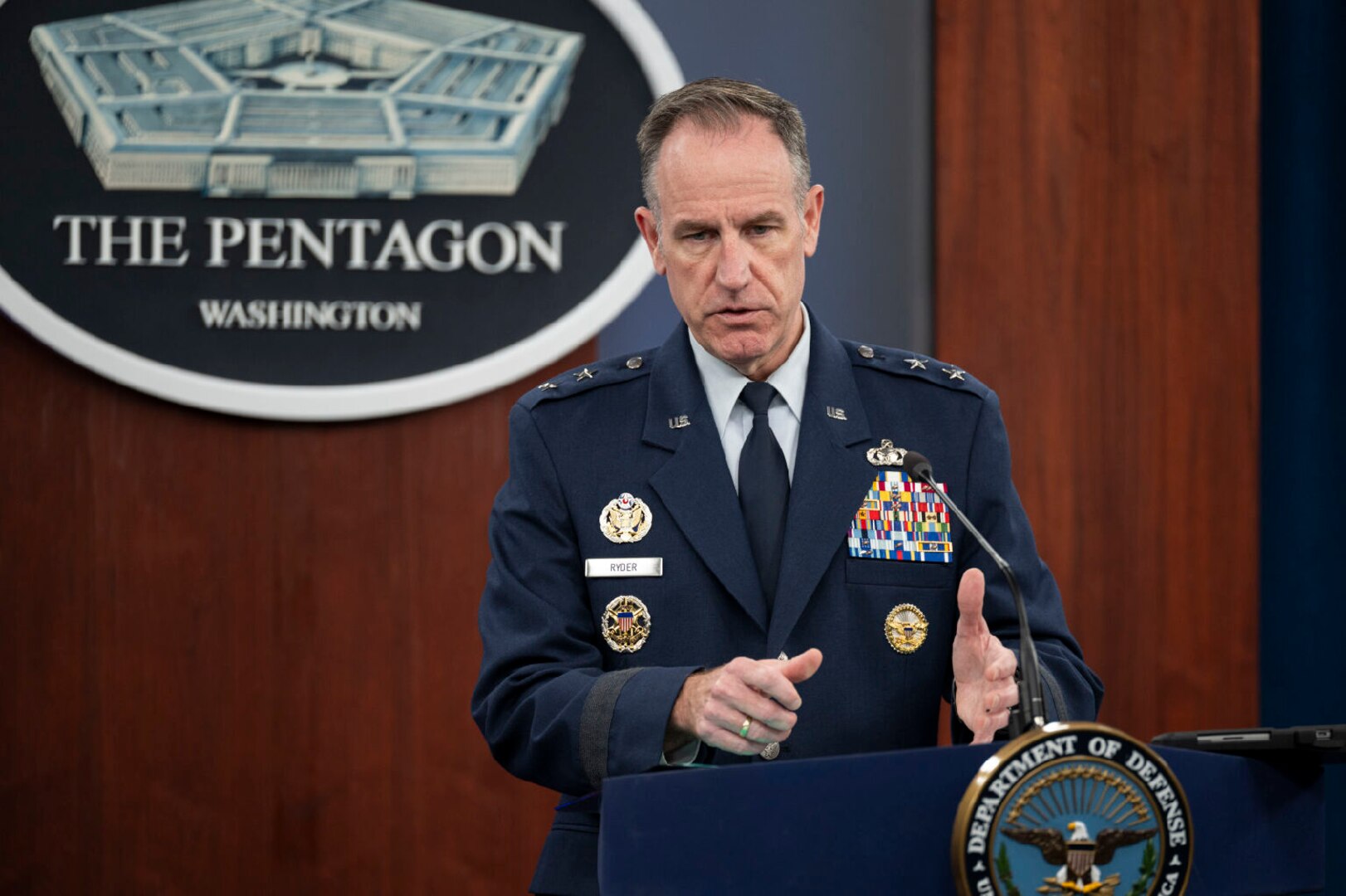 A man in a dress military uniform stands at a lectern talking and gesturing with his hands.