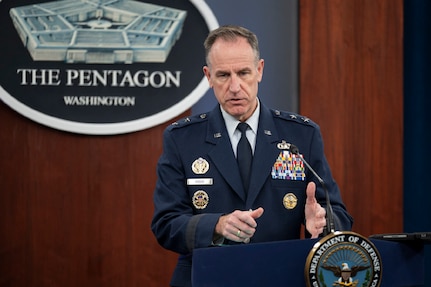 A man in a dress military uniform stands at a lectern talking and gesturing with his hands.