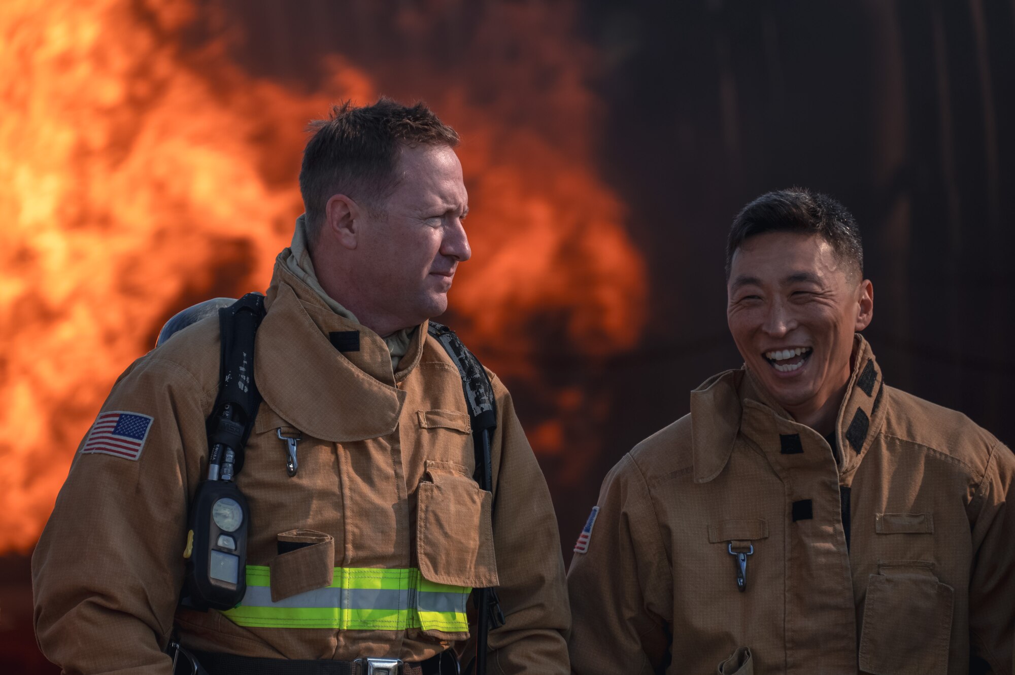Two men stand with fire in the background