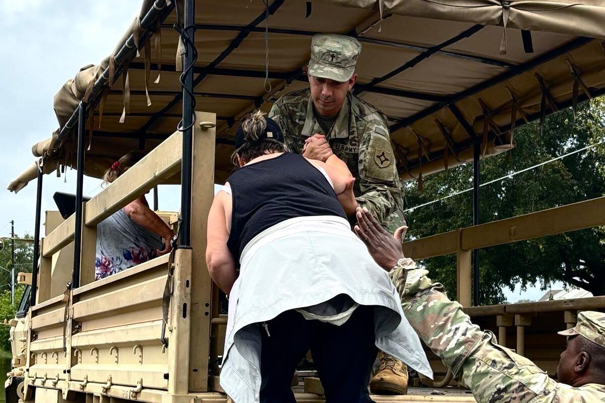 Two soldiers help a civilian climb up and into the open back of a military vehicle.