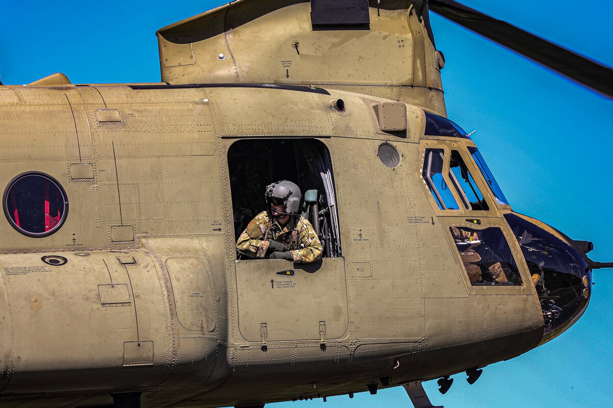 A soldier wearing a helmet and headset looks out from a helicopter as it hovers in the air against a blue sky.