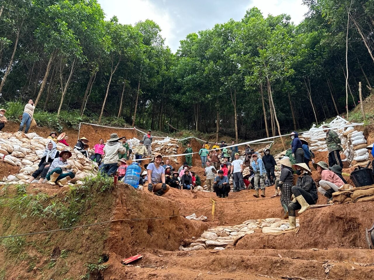 A group of people crouch on the ground and pose for a photo as they work on uneven ground in front of a forest.