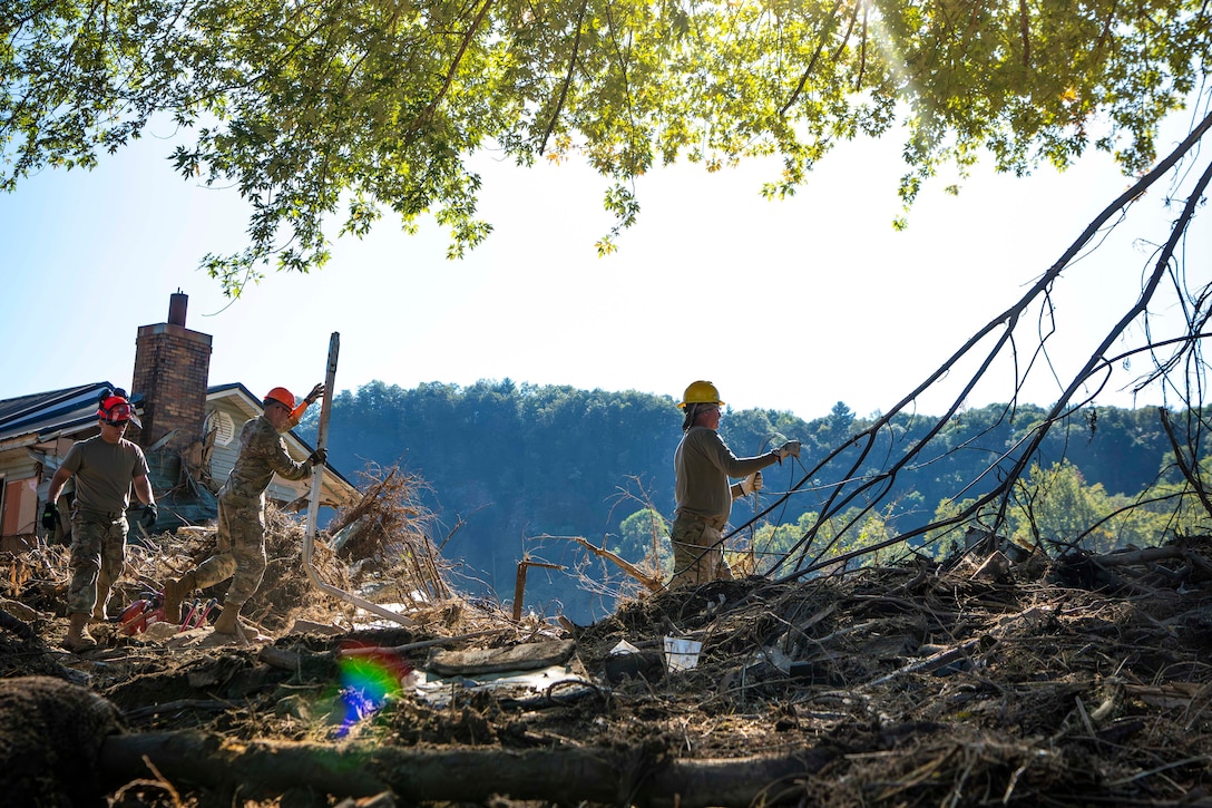 Three airmen wearing hardhats clear trees and brush near a house. A line of green trees is in the background.