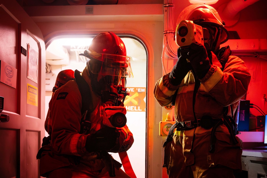 Two sailors wearing firefighting gear stand in a small room with red lighting on a ship. One holds a hose while the other holds a thermal imaging camera.