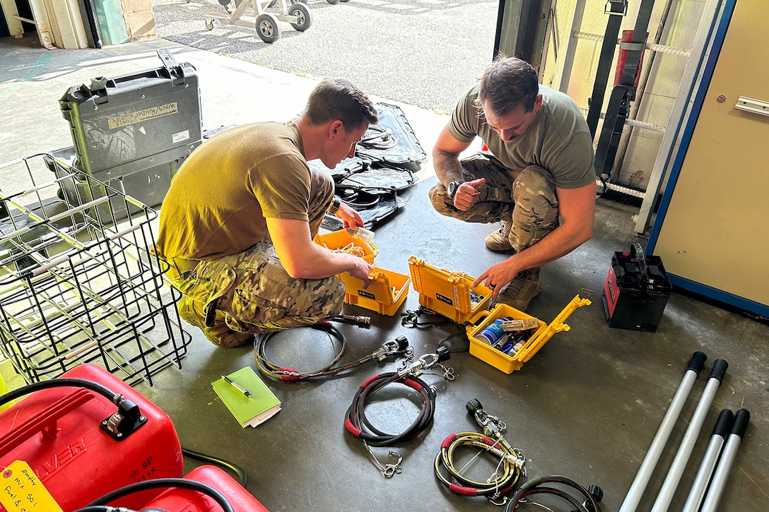 Two airmen squat to look through small containers on the ground as they prepare search and rescue equipment.