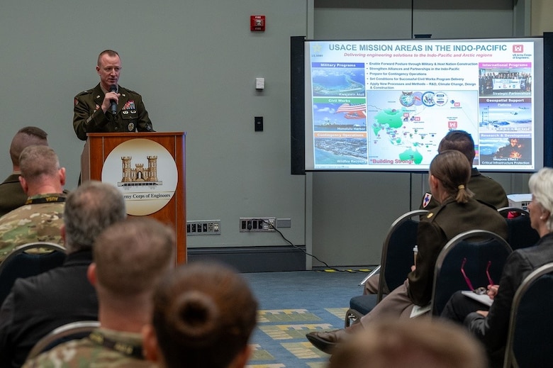 Soldiers sit in a room where a Soldier in uniform addresses them from a podium. There is a projector screen showing different mission areas USACE performs in the INDO-PACIFIC region in the front of the room