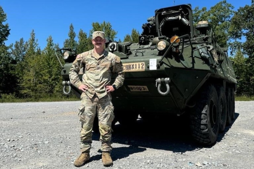 A smiling soldier stands with hands on hips in front of a military vehicle parked outdoors.