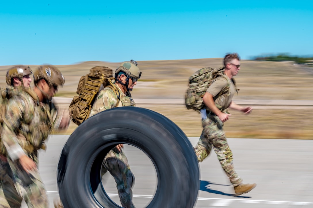 Airmen run while pushing a tire next to a field in the daylight.