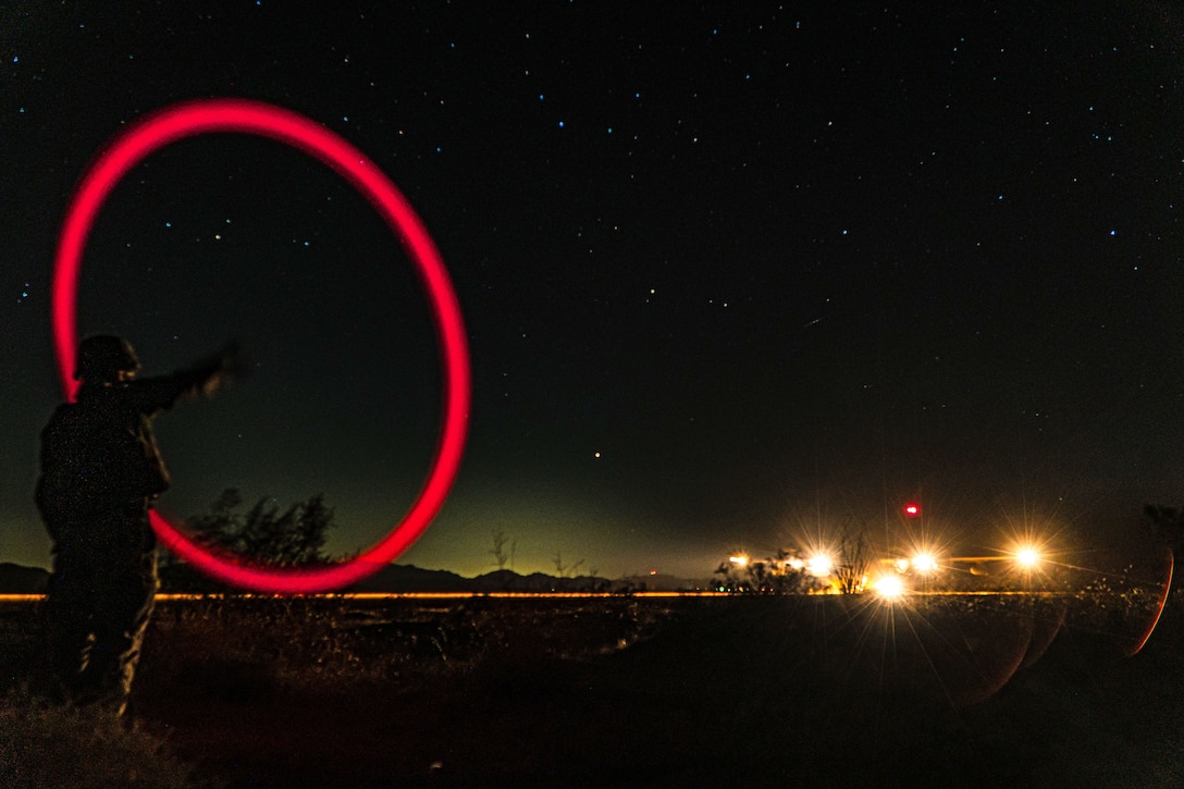 A sailor creates a red circular signal for an approaching aircraft shining its headlights under a starry night sky.