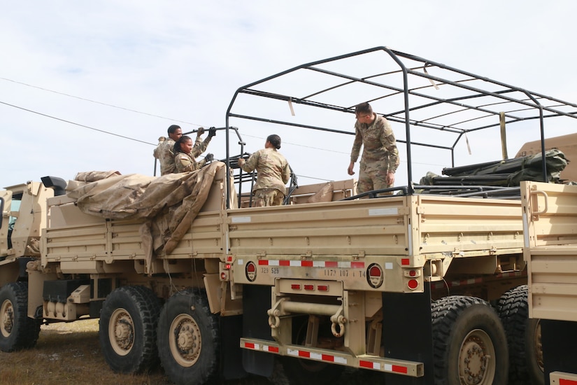 Soldiers adjust metal bars on top of the the open beds of two military trucks.