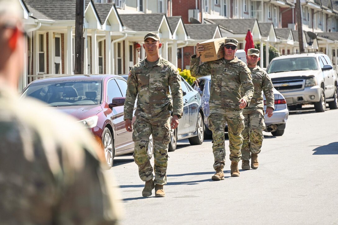 Three soldiers, two carrying boxes on their shoulders, walk down a residential street as a fourth, shown from behind, stands by.