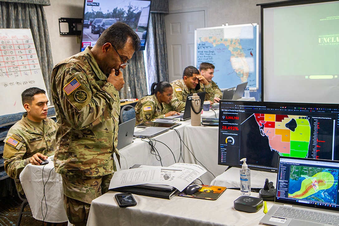 A standing soldier looks at documents on a table near monitors displaying storm data as other soldiers sit at laptops.