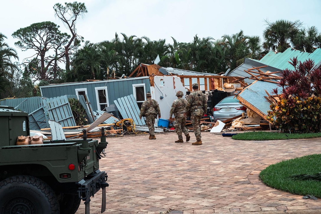 Three soldiers walk along brick hardscaping by a decimated bluish-gray wood-framed house.