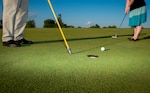 closeup of two sets of feet, a golf hole, ball and putter.