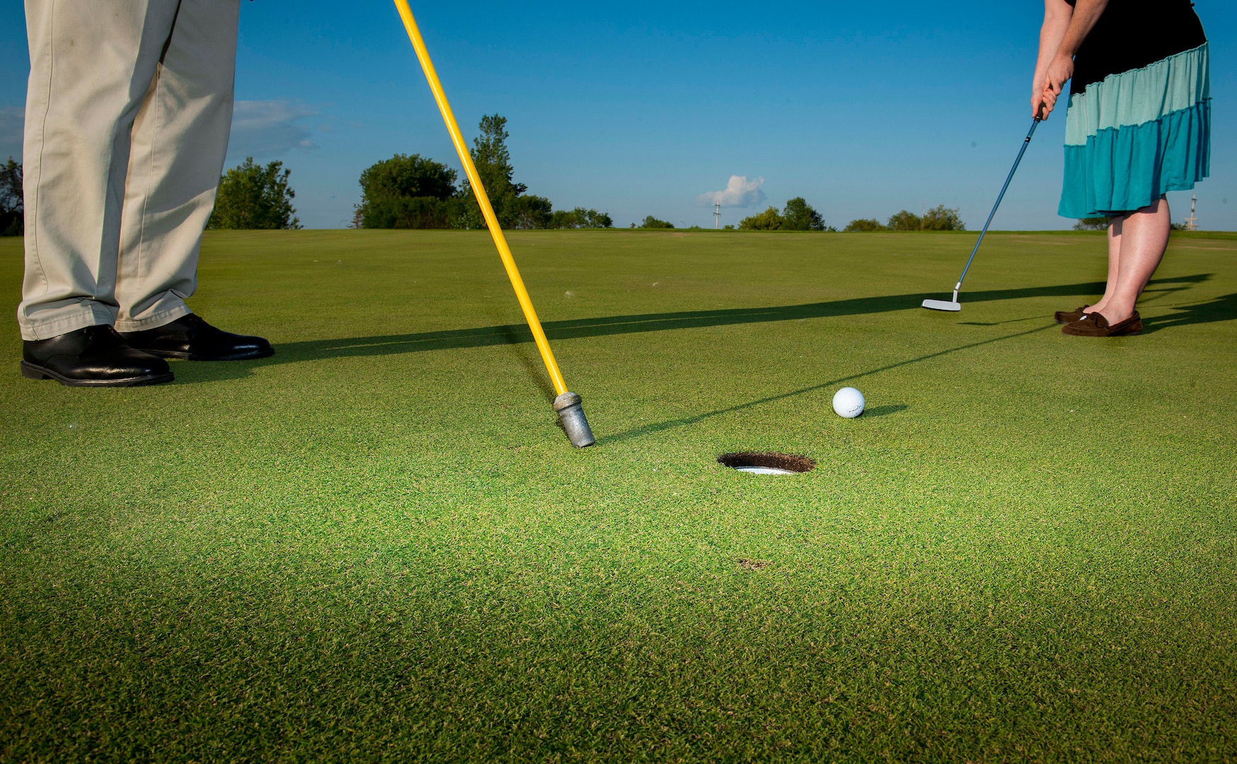 closeup of two sets of feet, a golf hole, ball and putter.