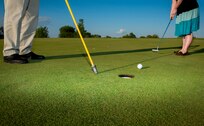 closeup of two sets of feet, a golf hole, ball and putter.