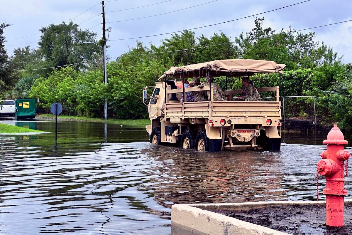 A truck with passengers moves over a flooded street on an overcast day.