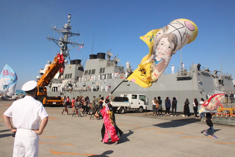 A Navy captain observes as USS Preble arrives in port while locals wave traditional flags.