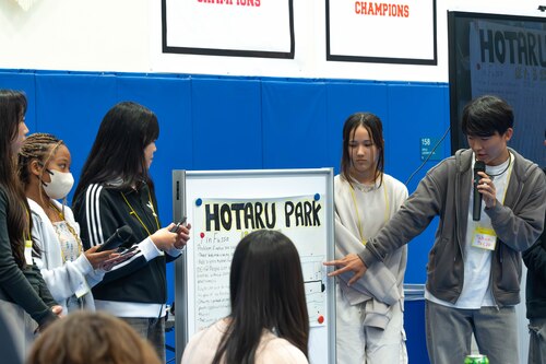 Student holding a microphone points to a board explaining his teams presentation.