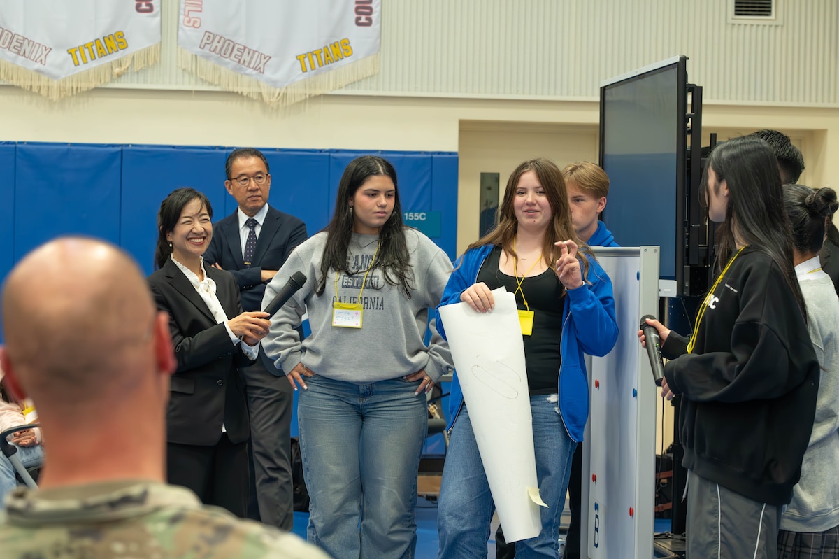 Student holds a white paper with one hand while speaking to a person sitting down.