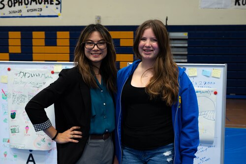 Teacher and student stand next to each other for a photo while standing in front of multiple white boards with the students presentations.