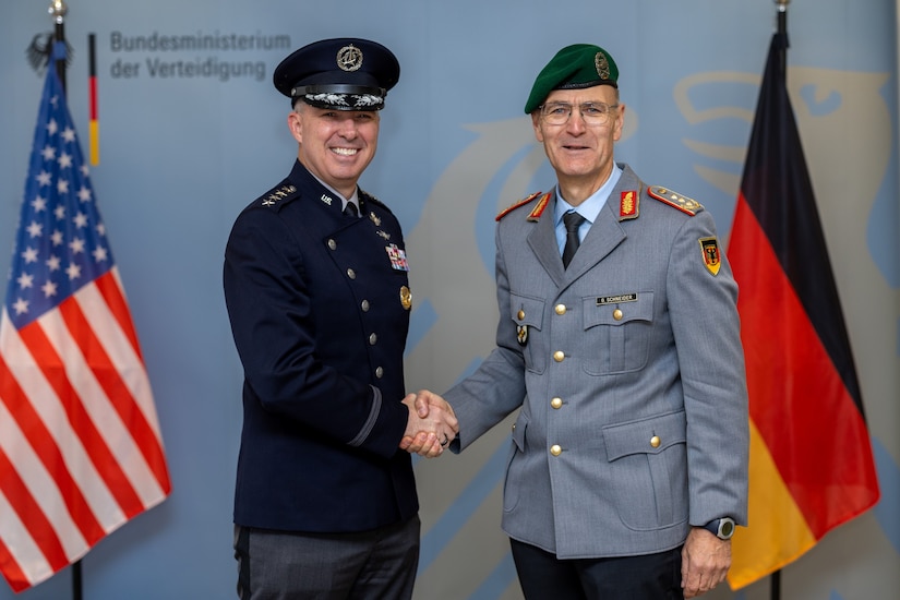 Two military officials in uniform shake hands in front of the German and American flags.