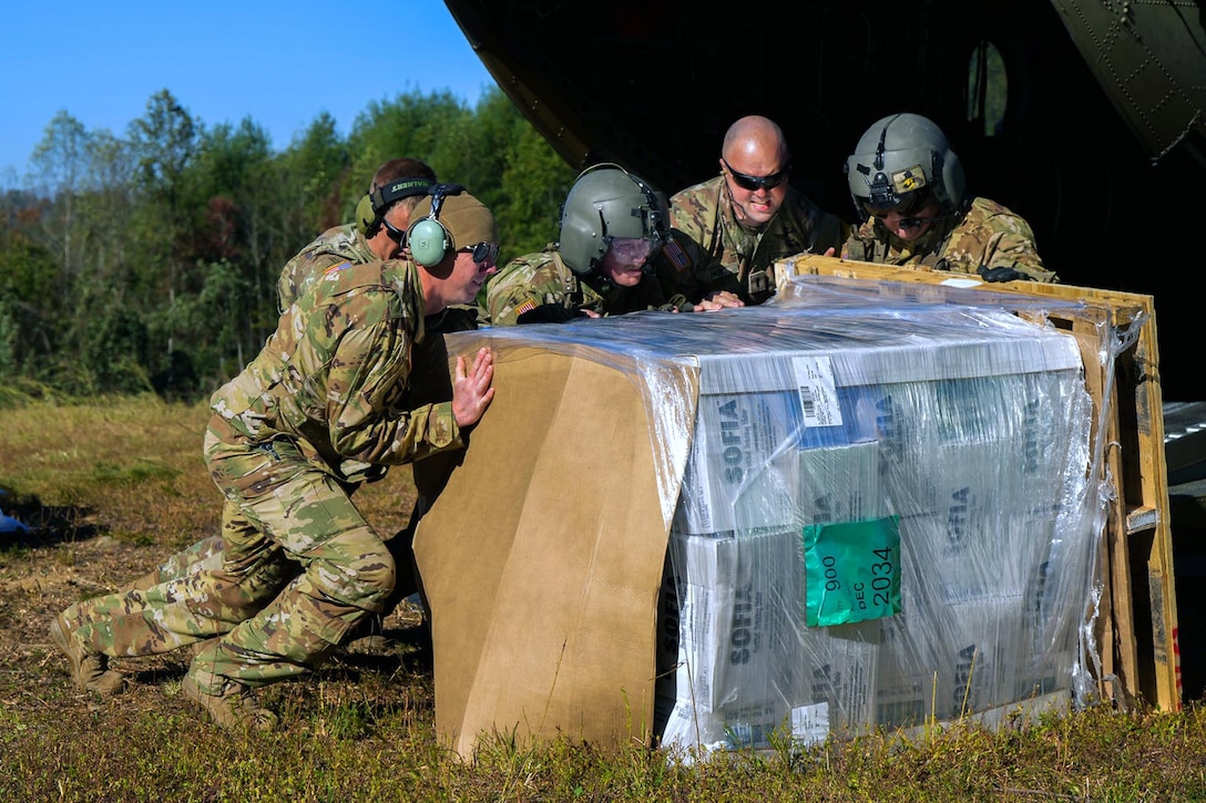 A group of soldiers push a large pallet holding essential supplies on the ground next to a helicopter.