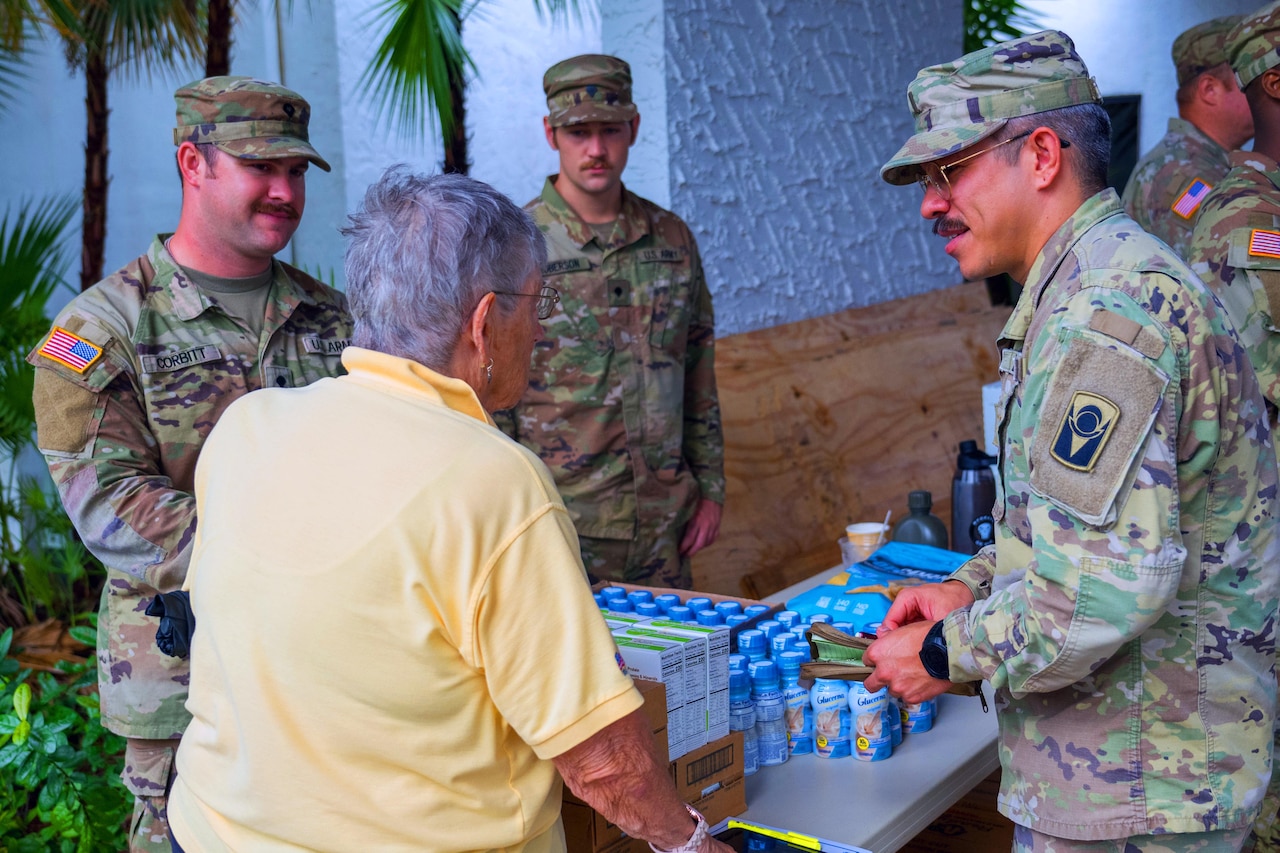A group of guardsmen chat with a resident by a table with essential supplies.