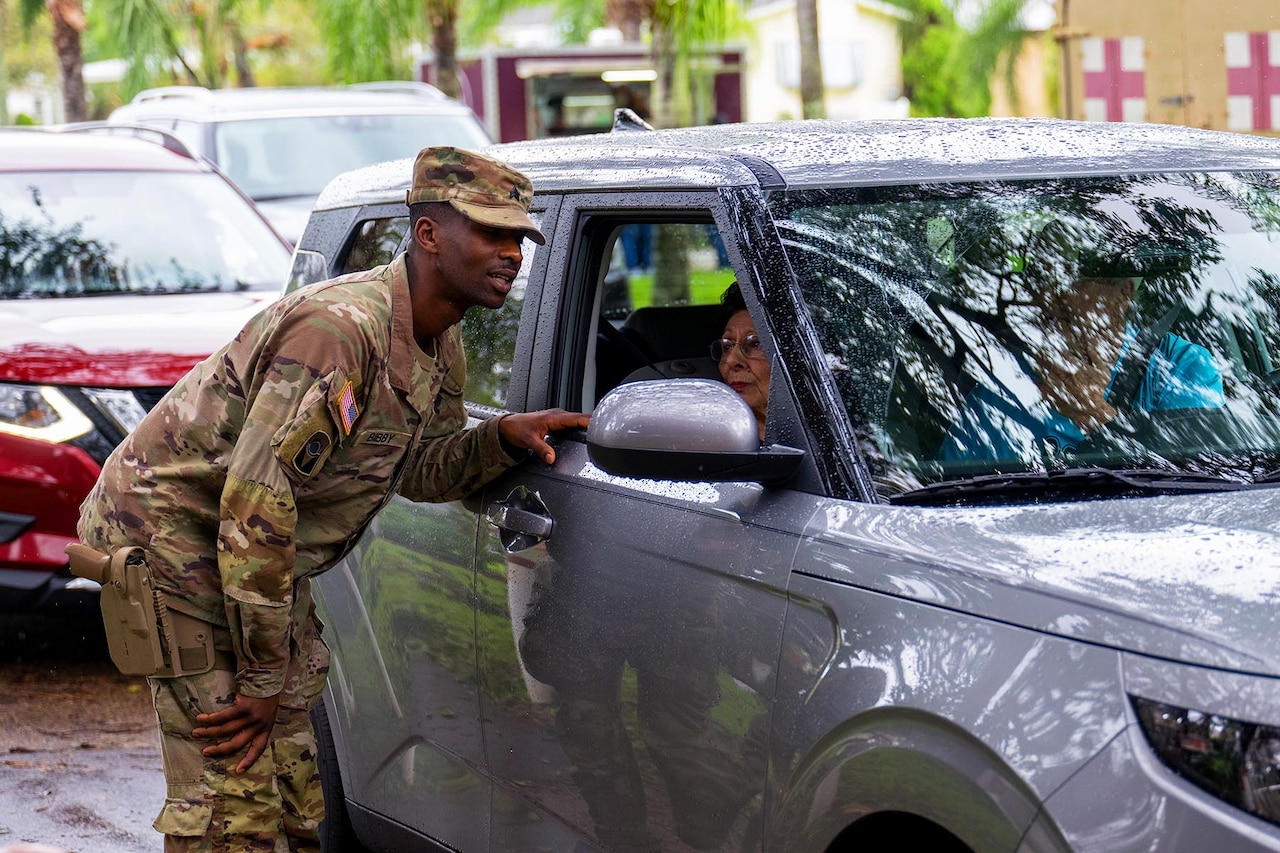 A guardsman chats with a passenger inside a grey vehicle during the day.
