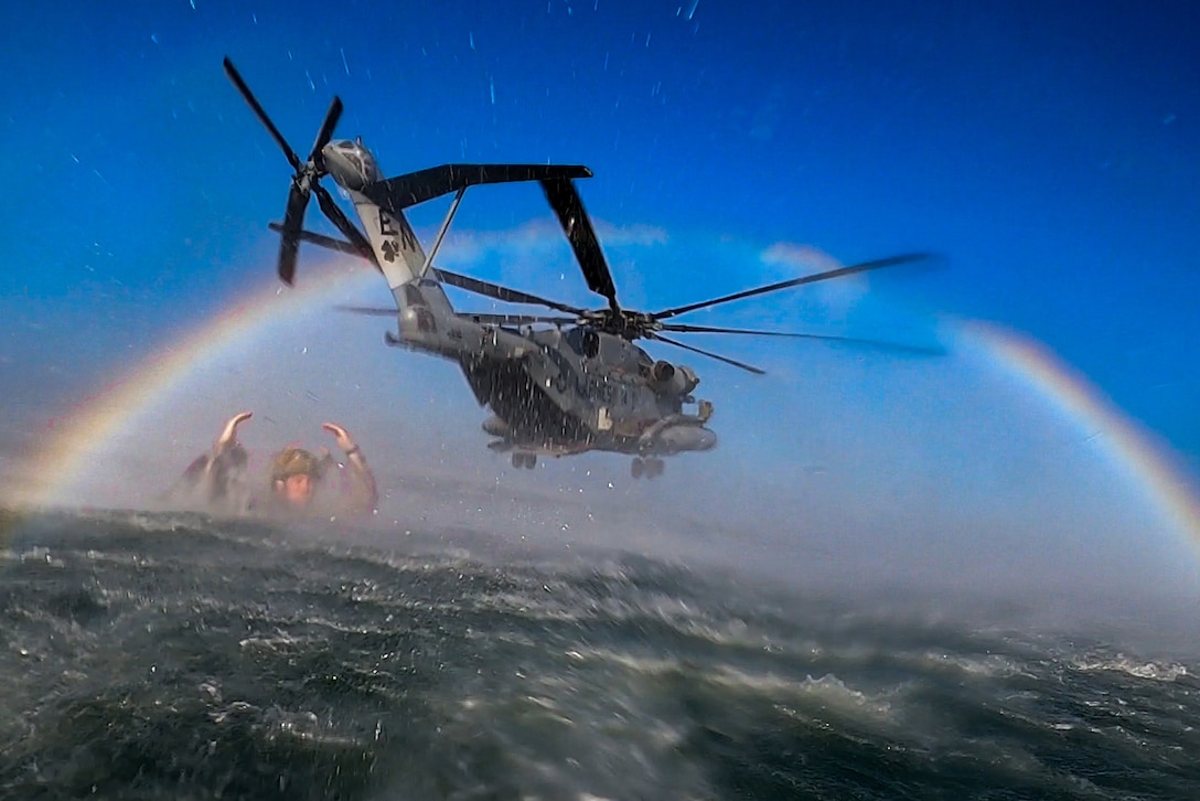 A Marine floats in a body of water as a helicopter hovers nearby under a rainbow.