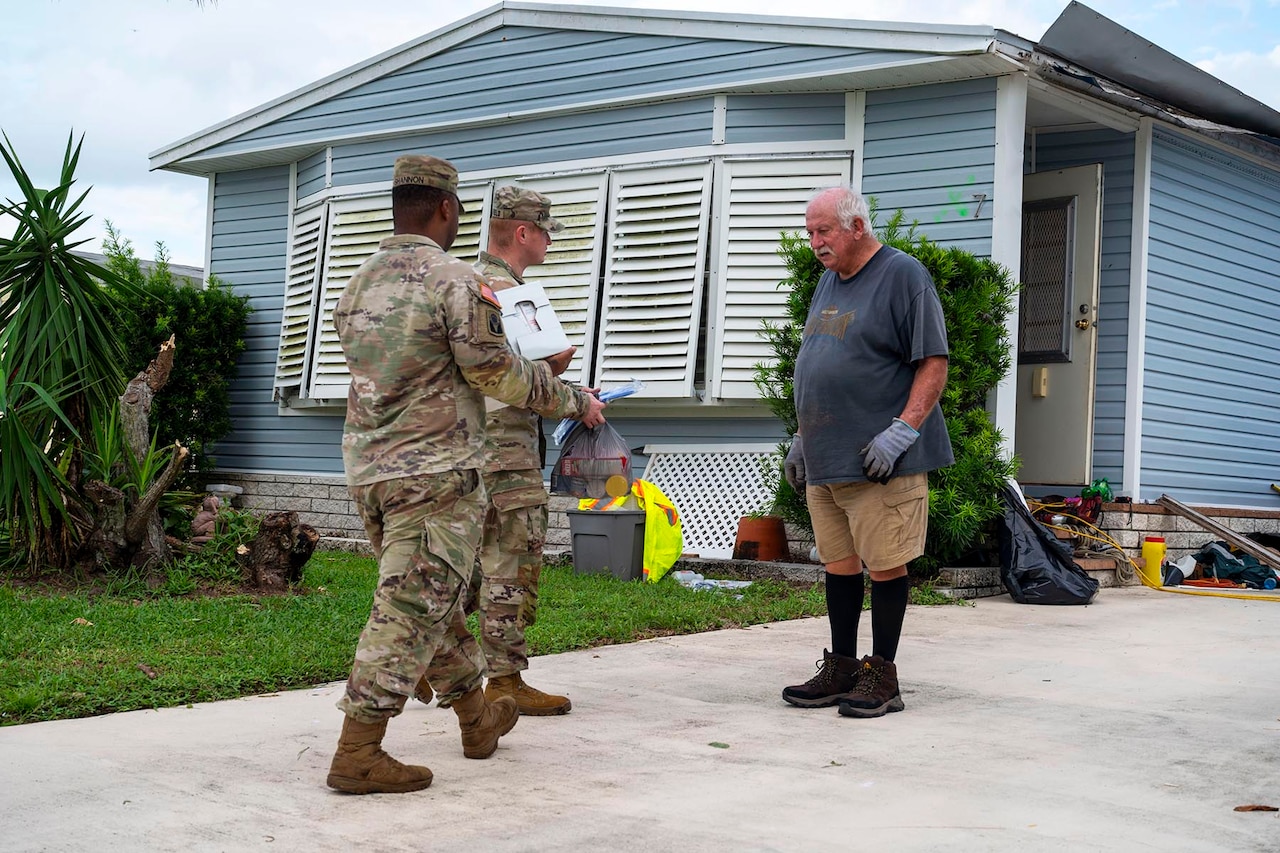 Two soldiers carry boxes and supplies to a resident standing outside their home.