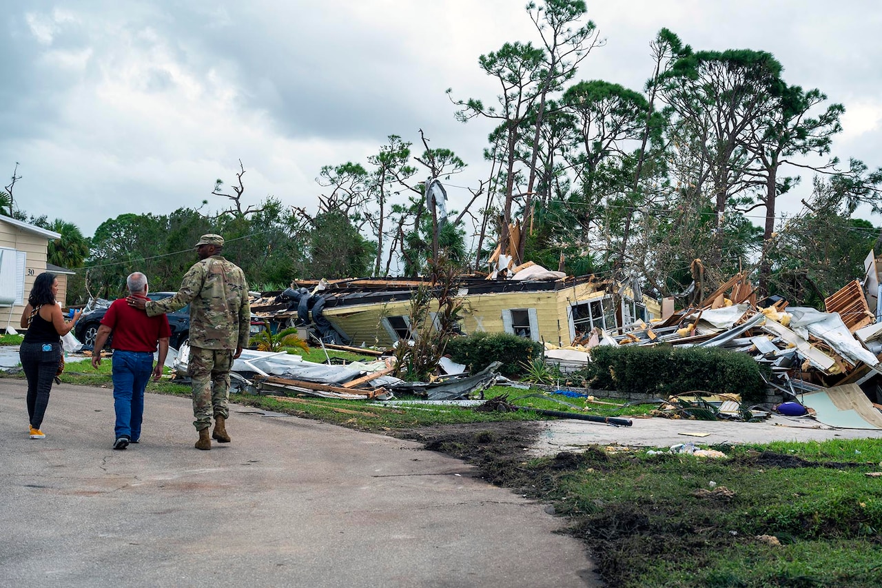 A soldier walks with two residents toward hurricane debris in front of a home.