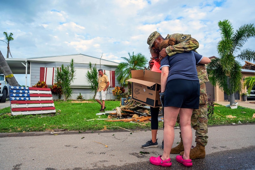 A soldier is hugged on either side by two residents while another looks on near hurricane debris in front of a home.