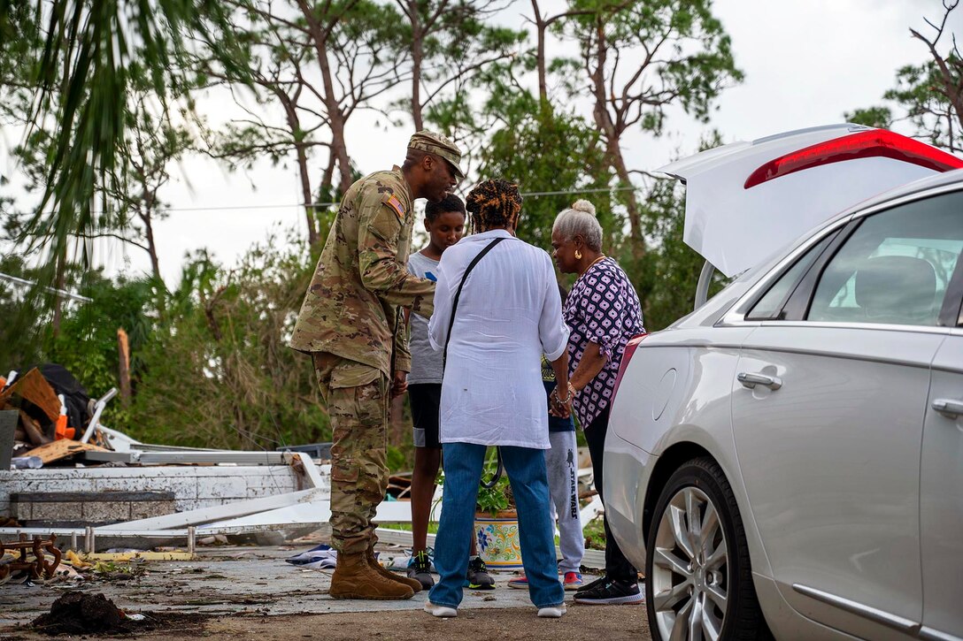 A soldier is surrounded by three residents behind a car with hurricane debris in the background.
