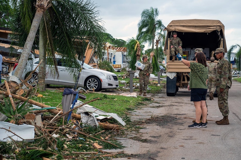 Soldiers stand outside a military vehicle with hurricane debris around while talking with a resident.