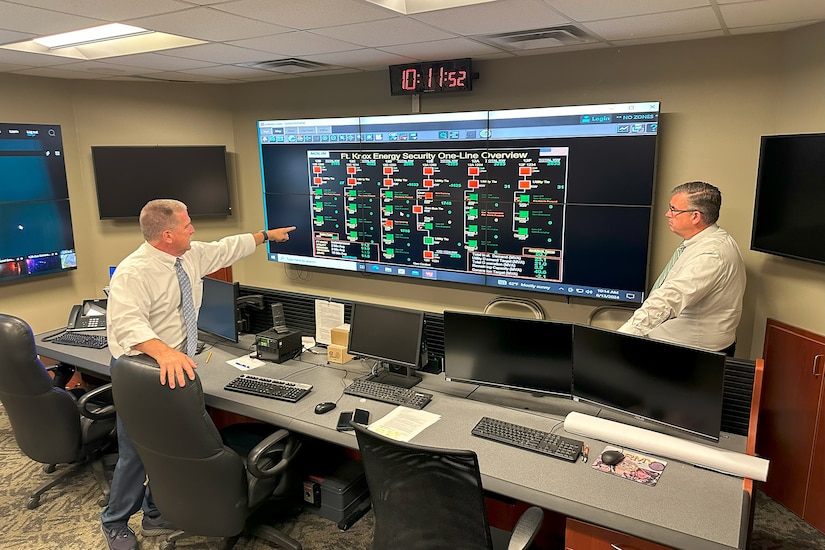 Two people in business attire look at a large digital board, with computers and desks nearby.