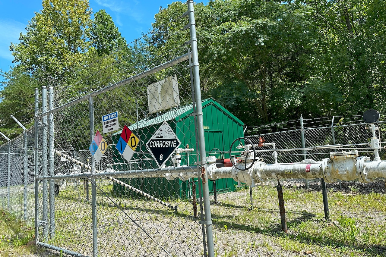 A small green building is surrounded by large pipes and a fence.
