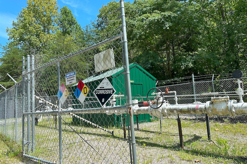 A small green building is surrounded by large pipes and a fence.