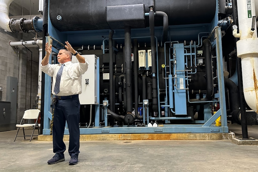 A person in business attire gestures in front of a large piece of equipment.