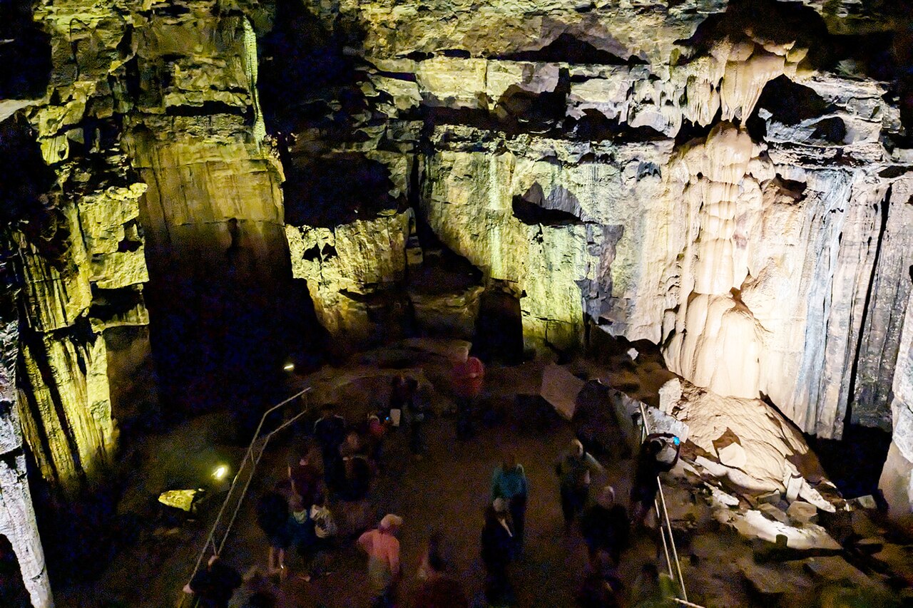 Lights shine on the walls of an underground cavern. Tourists mill about on the ground below.