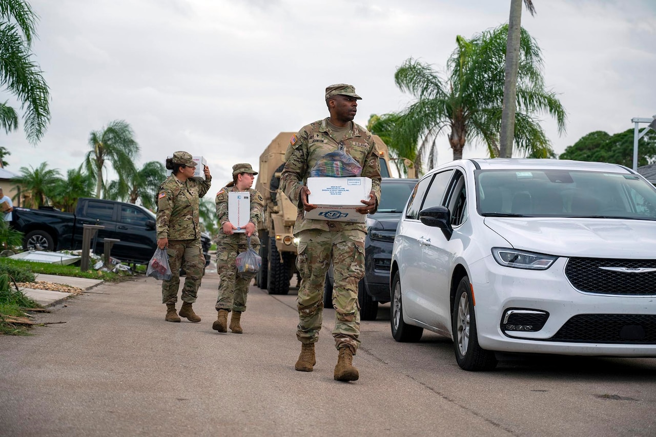 Three soldiers walk up a residential street next to civilian vehicles carrying care boxes.