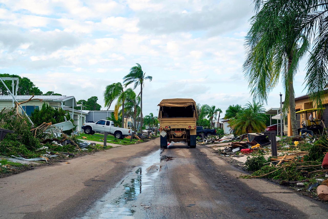 A military vehicle drives along a residential street with hurricane debris on either side.