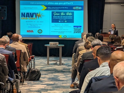 A woman stands behind a podium in front of a full seating area, presenting a slide deck at the Small Business Symposium during Carderock's Annual Industry Day.