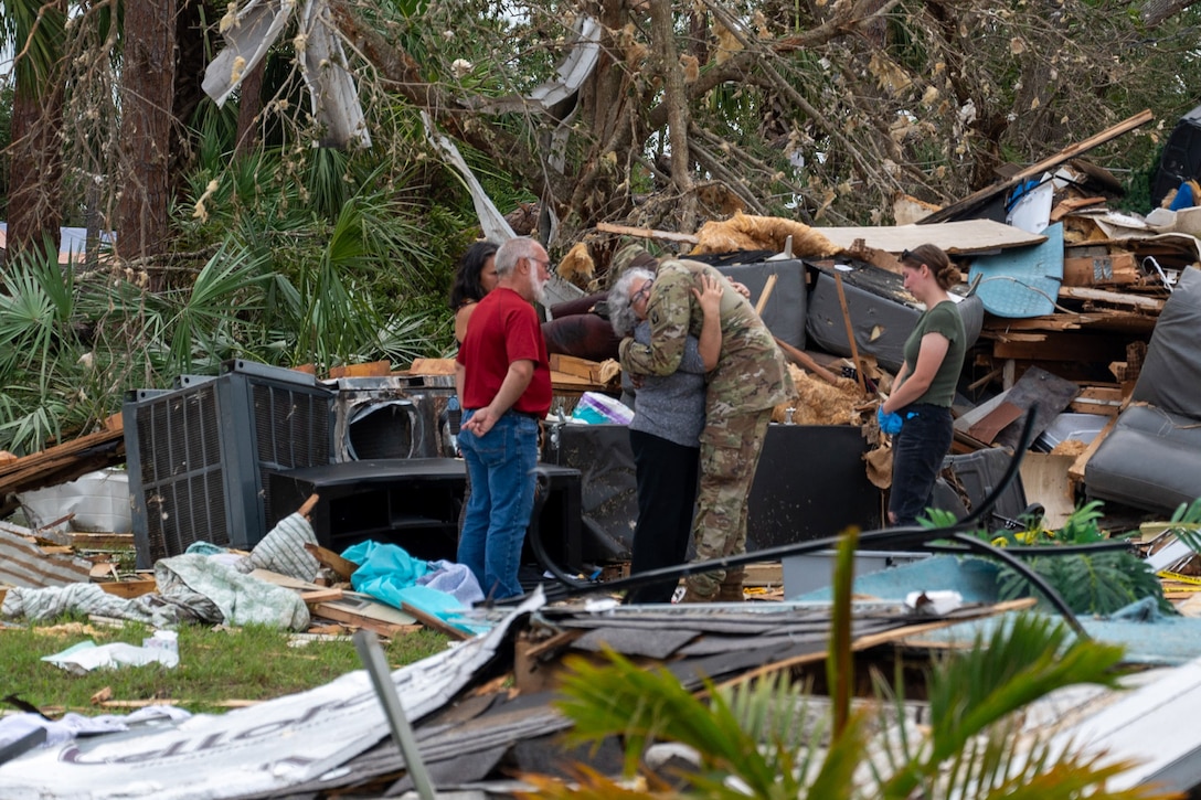 A soldier hugs a civilian and three other people look on, while they all stand in front of a huge pile of wreckage and storm debris.