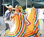 A man in a grey suit and sombrero and a woman in a colorful yellow dress with rainbow stripes are dancing a traditional Hispanic dance