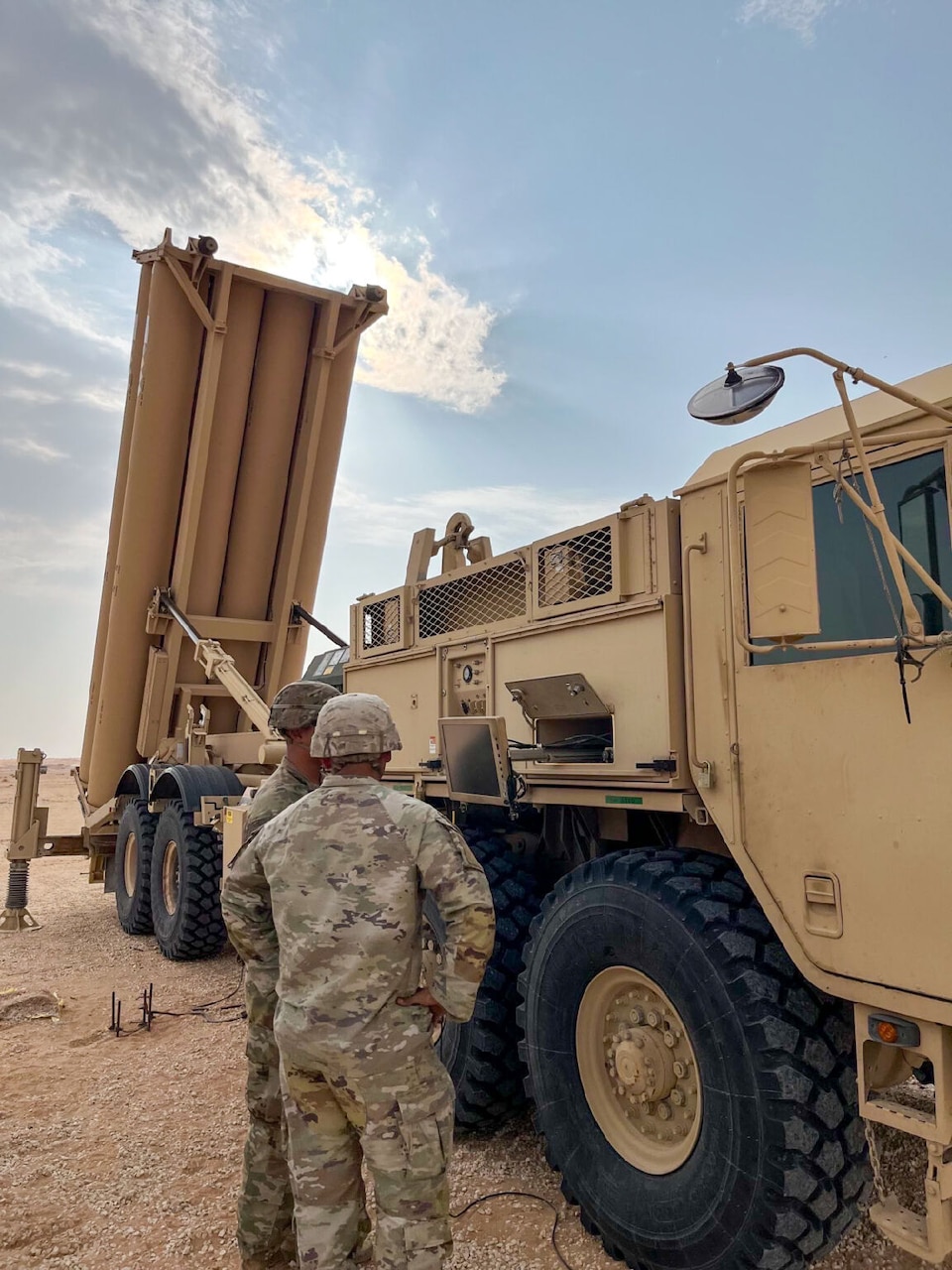 A man in camouflage fatigues with his back to the camera and another man dressed the same way are talking next to a large military truck with a vertical launch tube attached to the back that is facing upward.