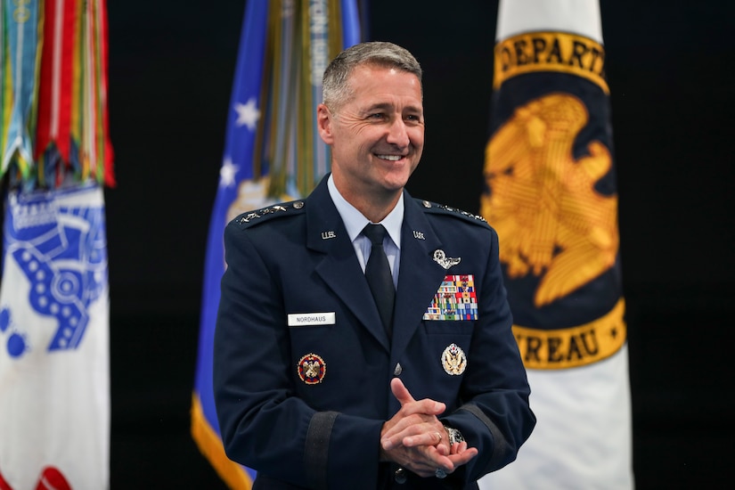 A man in military uniform smiles in front of three flags.