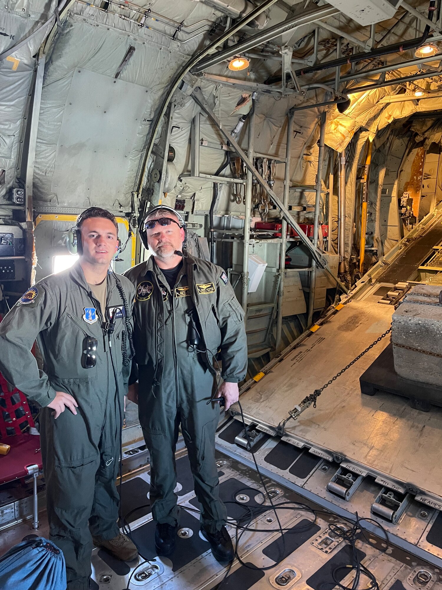 A U.S. Air National Guard loadmaster and a civilian contractor for the U.S. Navy pose for a photo arm-in-arm inside the back of a KC-130T aircraft near the rear ramp door.
